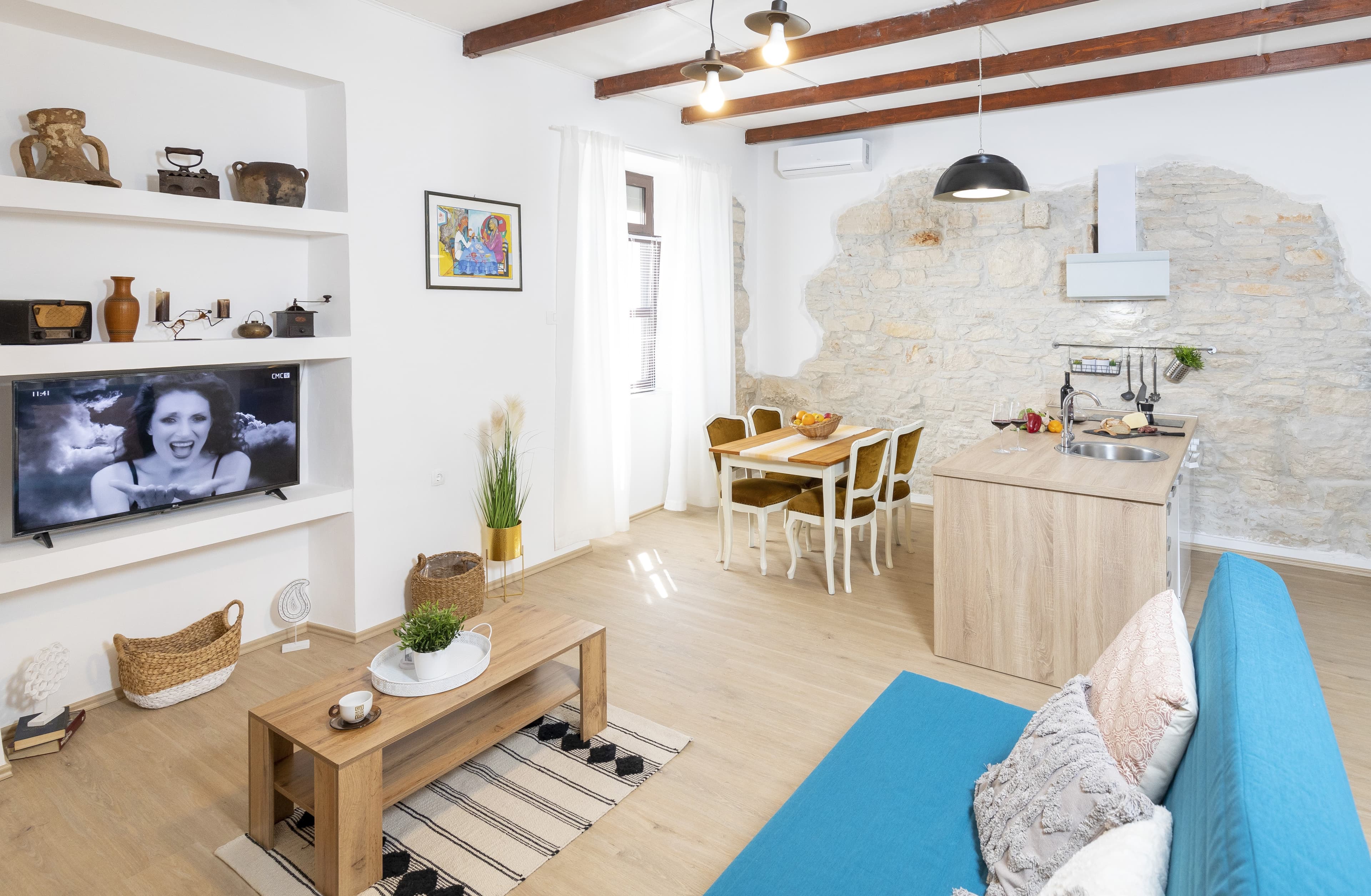 Modern open-plan living space with a blue sofa, stone wall, and rustic ceiling beams.