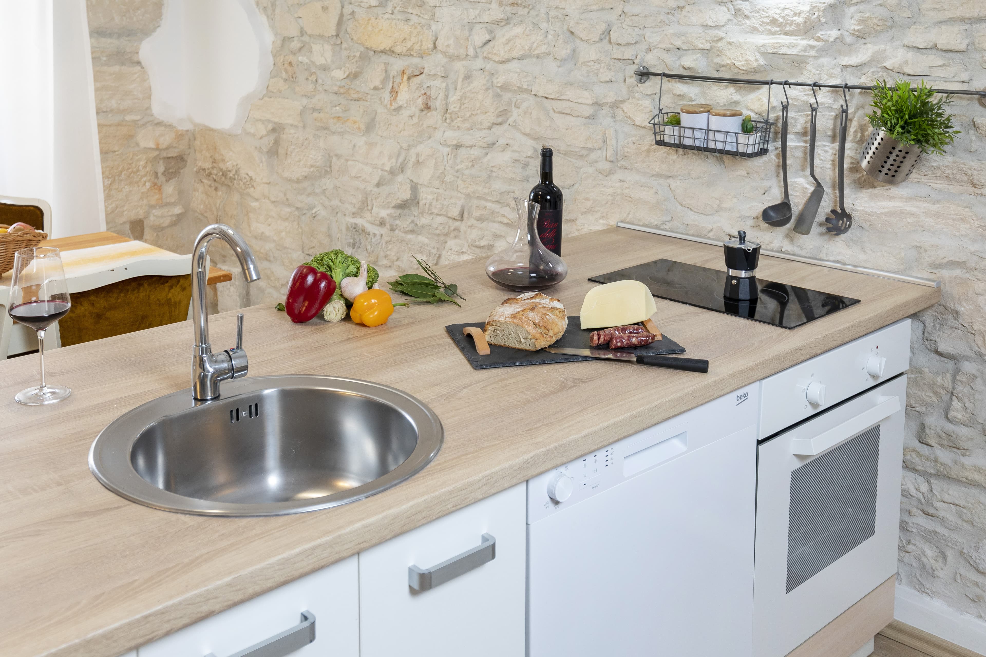 Rustic kitchen counter with bread, cheese, wine, and vegetables against a stone wall.