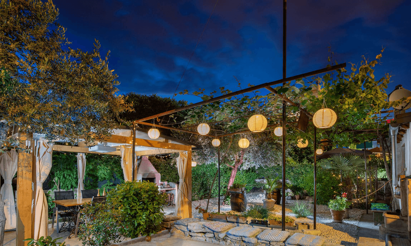 Glowing paper lanterns illuminate a cozy garden patio with a wooden pergola and lush greenery.